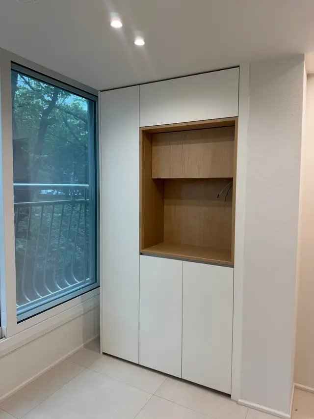 창가 옆 천장까지 닿는 흰색 수납장 가운데에 오크색 열린 칸이 두 단 있다.